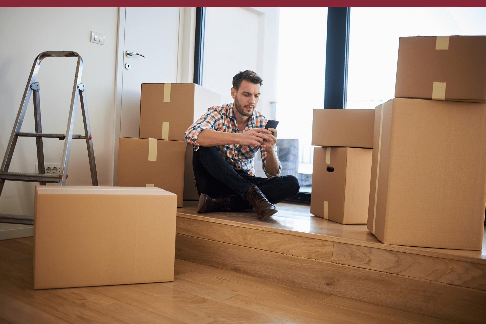 Man sitting on a wooden floor surrounded by cardboard boxes, looking at his phone during a move. A metal ladder is nearby, and natural light comes in through large windows behind him.