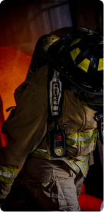 Firefighter in full gear walking towards the camera, bent over as if pulling a weight. The glow of a fire is visible behind him
