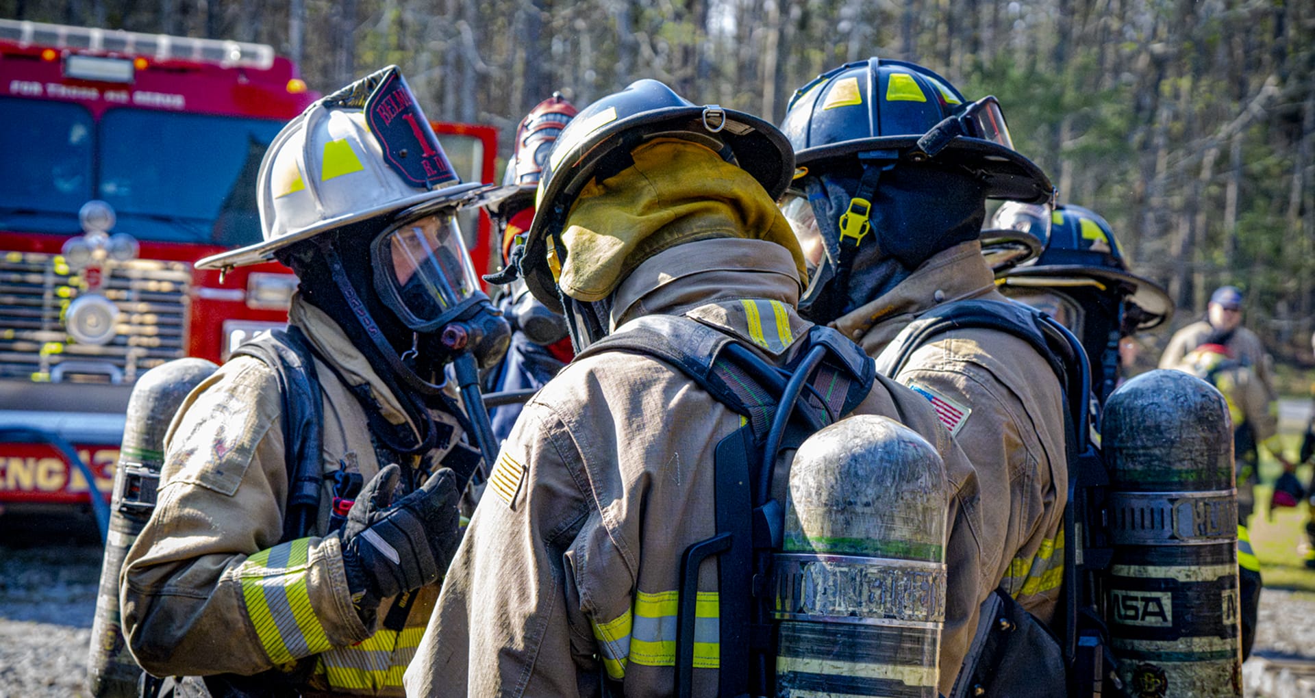 Several firefighters in full gear outside near some woods, huddled close and talking. A fire engine on a gravel area visible behind them