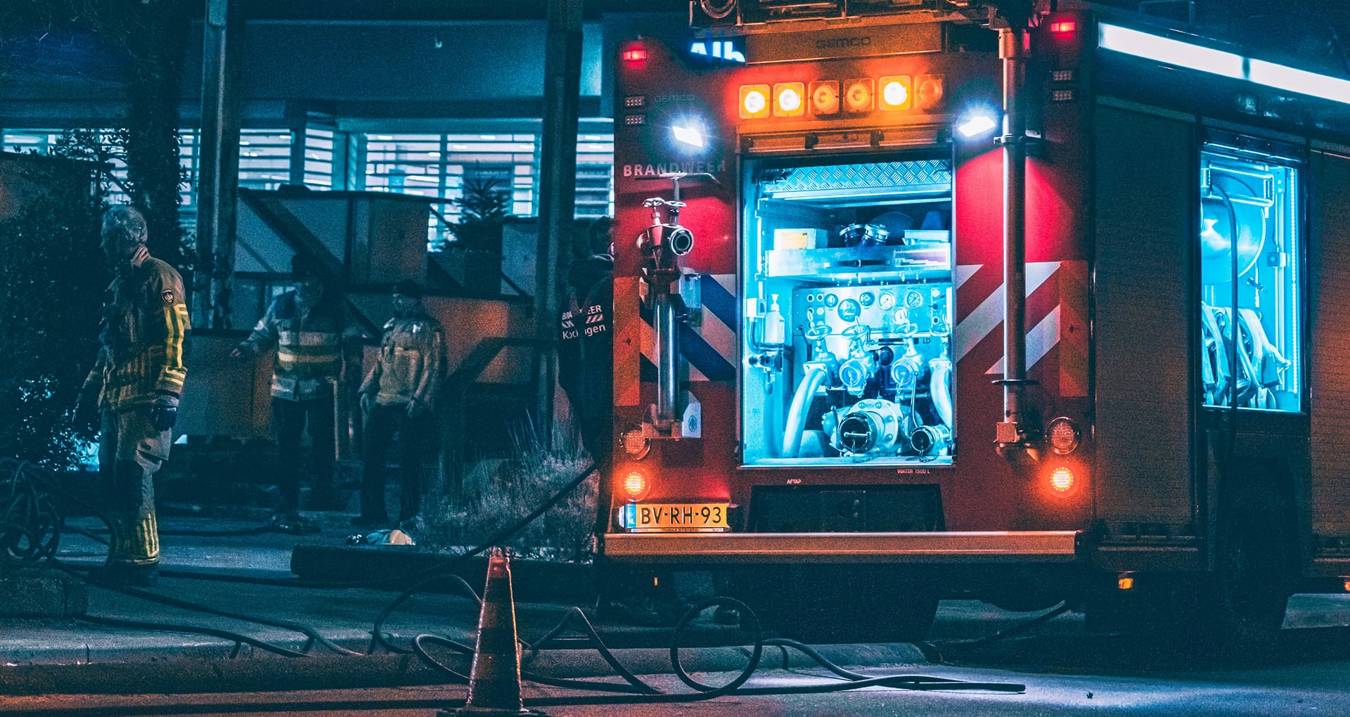 A fire engine at night, with its lights on illuminating the sidewalk and three firefighters working next to it, with a small hose on the ground