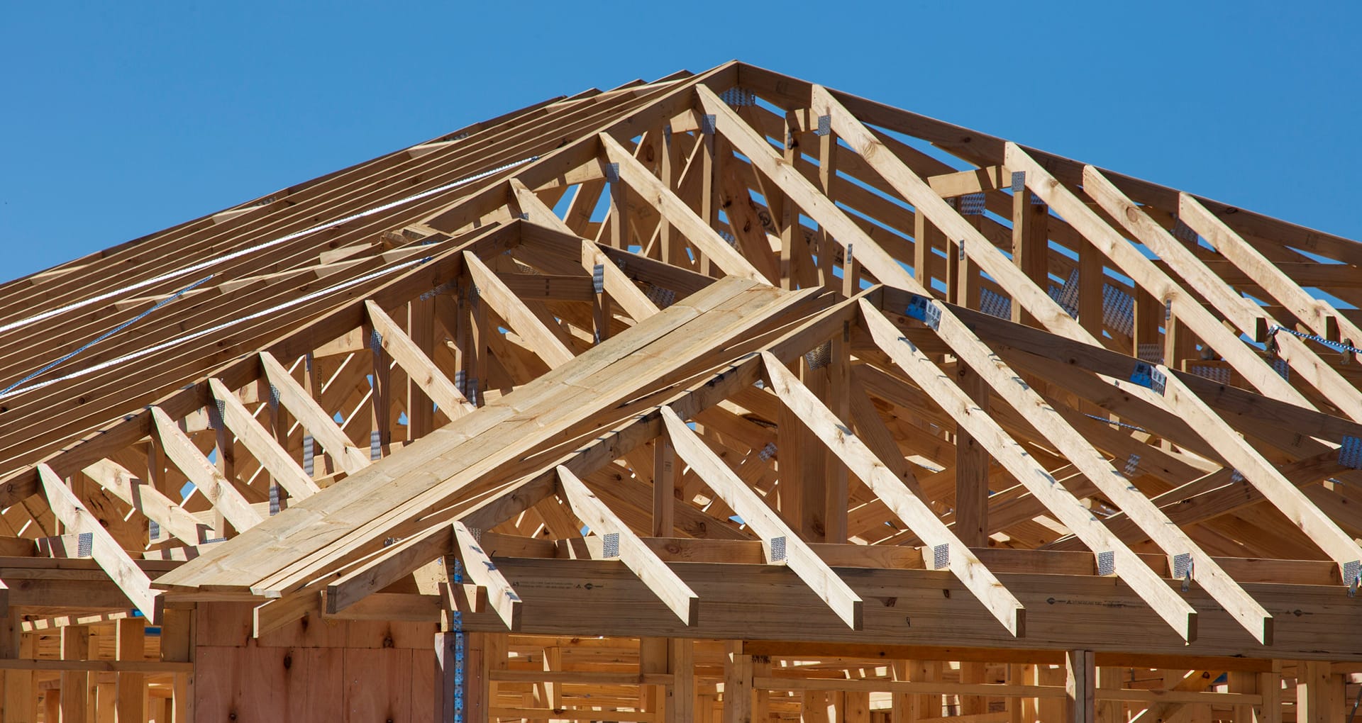 A photo of a building under construction, all wood framing, showing the roof trusses and a blue sky.