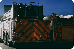 Two fire engines parked next to each other outside at dusk, viewed from behind, with a dark blue sky visible above, and a harsh light illuminating them from the side.