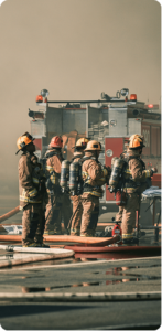 Five firefighters in full gear behind a fire engine, with hoses arranged at their feet and foreground, heavy smoke in the background.