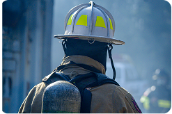 A firefighter in full gear, facing away from the camera, with smoke obscuring what he is looking at.