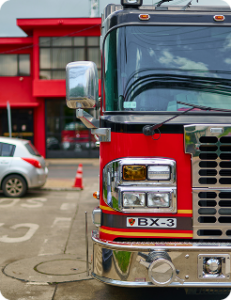 A photo of the front of a fire engine, in a parking lot across the street from a red two story fire station. A second fire engine is visible in the bay of the fire station