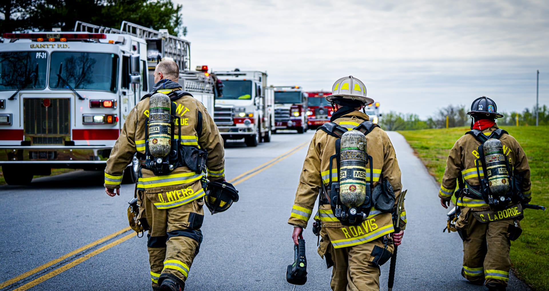 Three firefighters in full gear walking away from the camera on a road, with four firetrucks facing them in the opposite lane, and a field on their right.