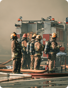 Five firefighters in full gear behind a fire engine, with hoses arranged at their feet and foreground, heavy smoke in the background.