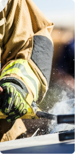 A closeup of a firefighter using a tool to break into what appears to be a damaged car. Only his gloved hand and some of his shoulder visible