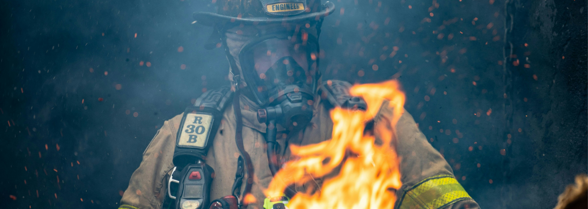 Firefighter engineer in full gear and SCBA mask surrounded by smoke and embers during an active fire