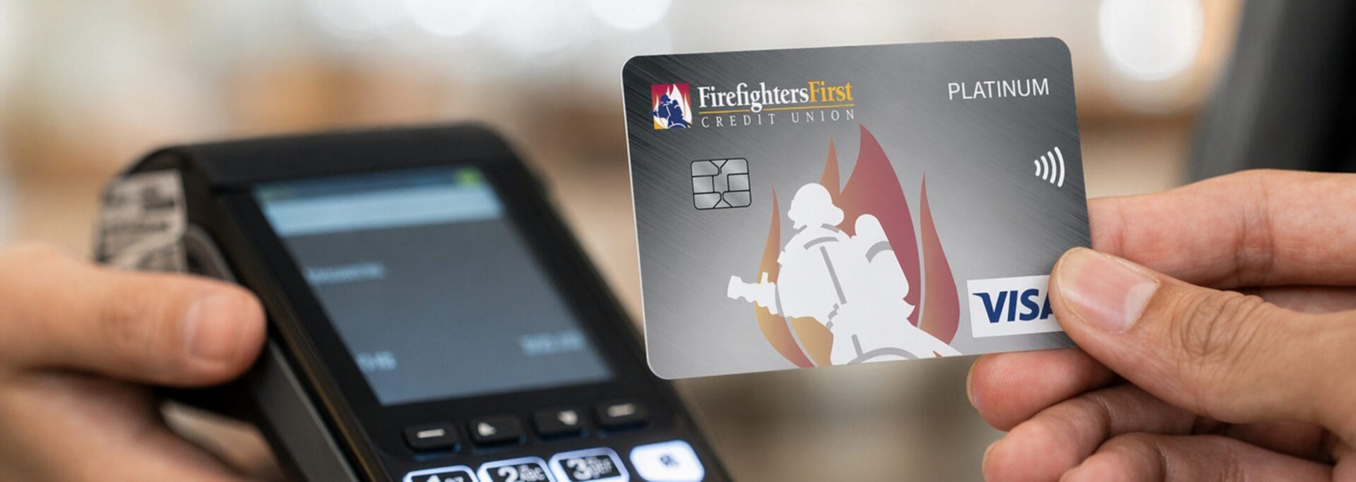 Hand holding a Firefighters First Credit Union Platinum Visa credit card in front of a payment terminal
