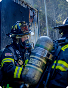Two firefighters in full gear with SCBA tanks working together at the scene of a fire-damaged structure