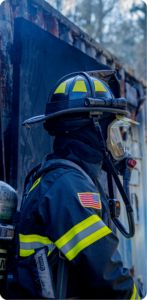 Firefighter in full turnout gear with SCBA and American flag patch standing near a fire-damaged structure