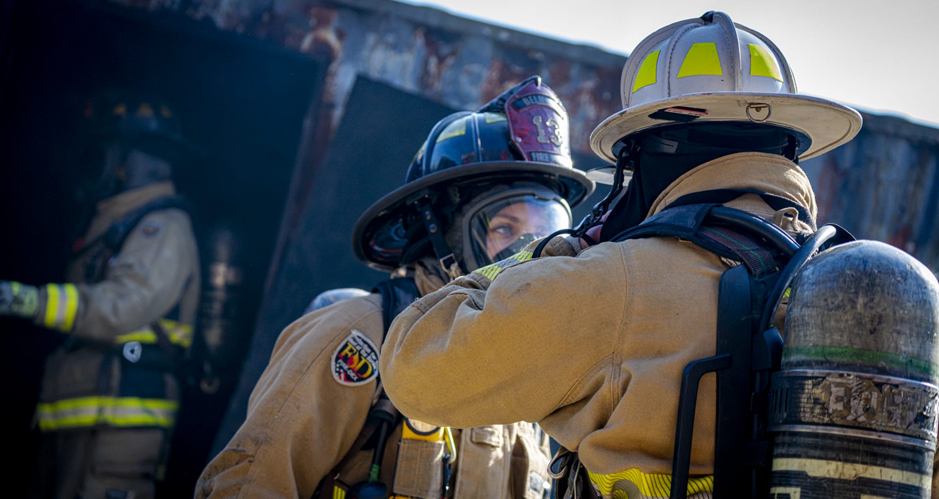 Two firefighters in full kit and oxygen mask talking to each other, with what appears to be a train behind them.