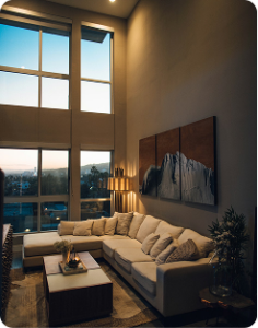 Cozy living room with a sectional sofa, tall windows, a floor lamp, and landscape artwork on a high wall.