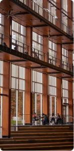 Multi-story building with large glass windows and wooden beams, with people seated on a terrace outside.