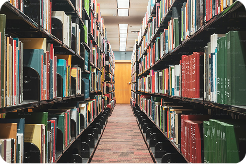 Narrow library aisle with tall bookshelves on both sides filled with books, leading toward a bright opening at the end.
