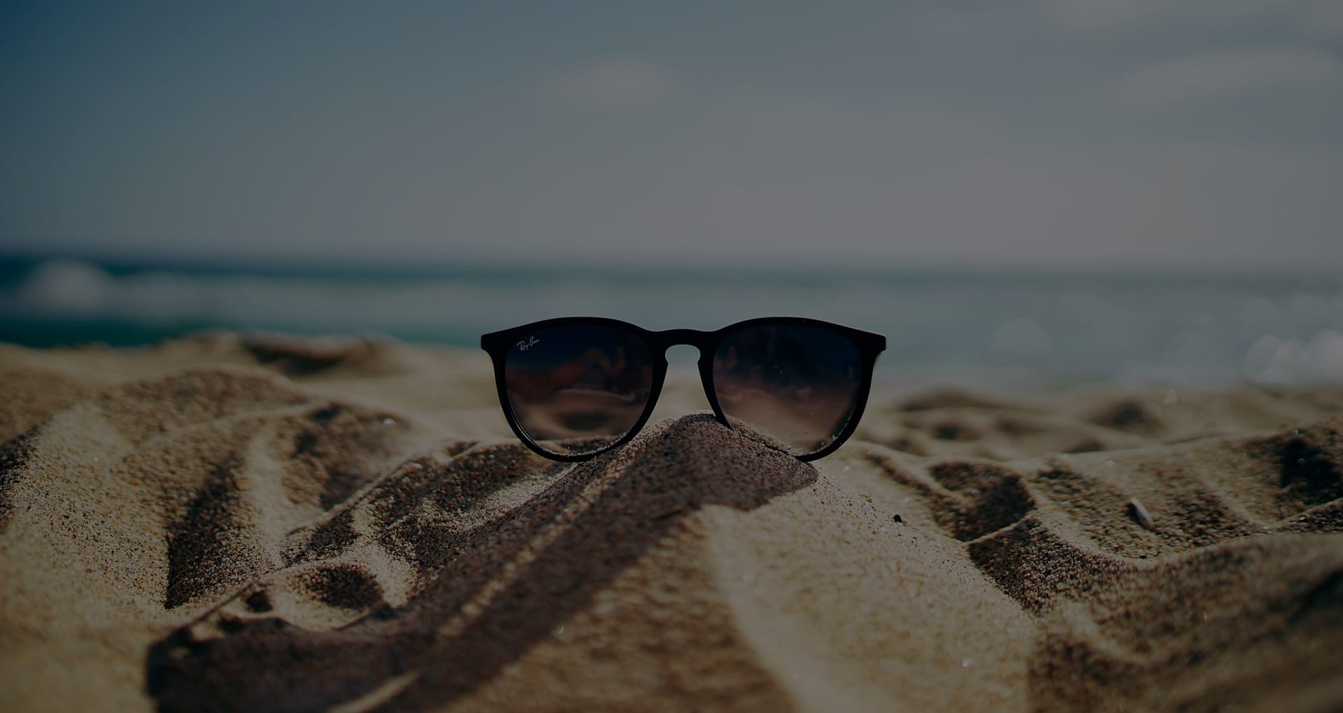 Sunglasses resting on sand at the beach with the ocean and sky blurred in the background.