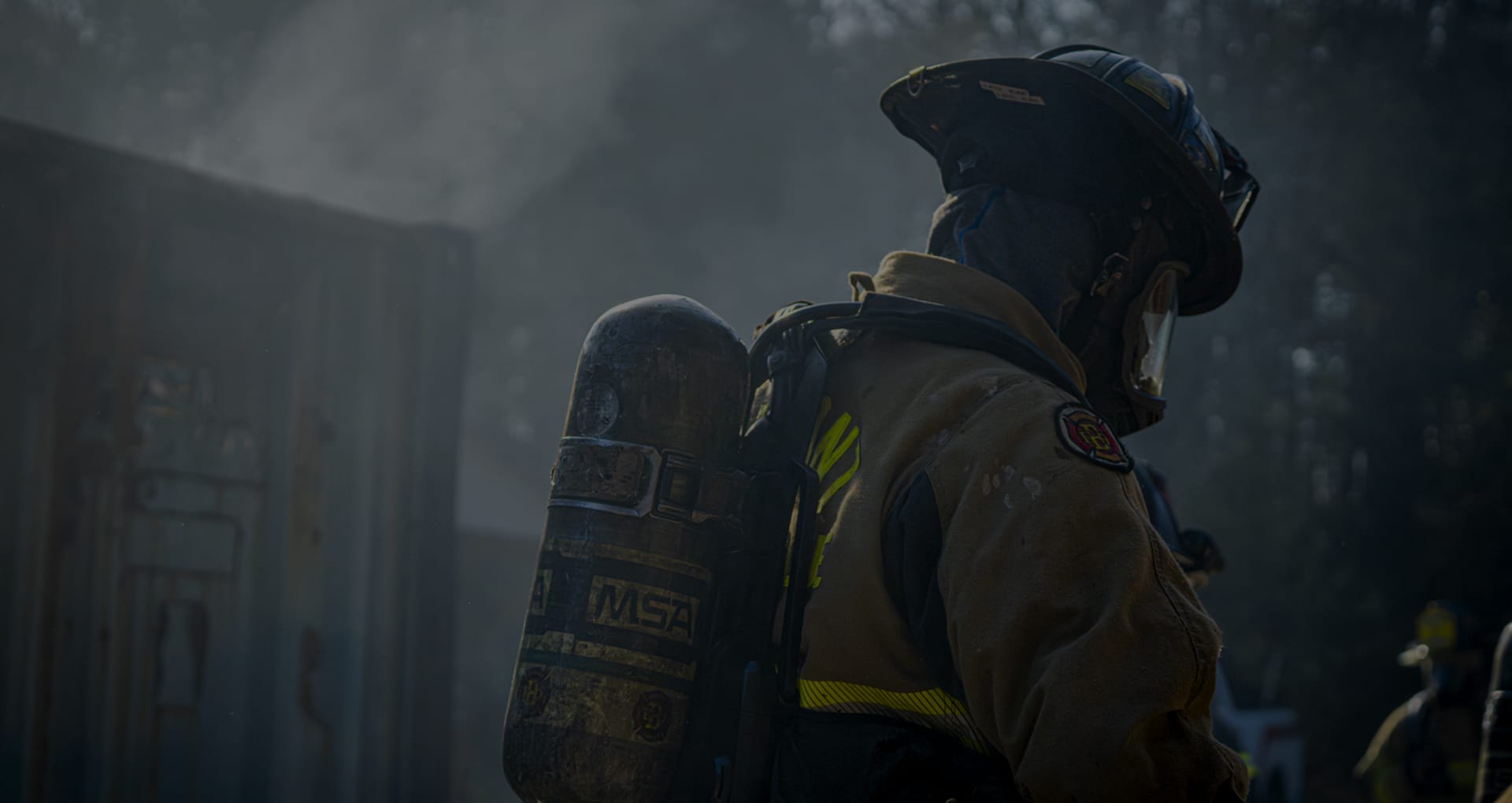 Firefighter in full protective gear with a helmet and face shield stands in a smoky outdoor scene, wearing an air tank on their back. Soft light filters through the haze, with trees and a building faintly visible in the background.