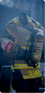 Firefighter wearing full turnout gear adjusts head protection with both hands while standing outdoors. The scene is lightly hazy, with sunlight filtering through and a structure partially visible in the background.