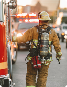 Firefighter in full gear walks beside a fire engine on a busy street, holding equipment with an air tank on their back.