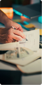 A woman's hand holding a pen, using it to review a document on a piece of paper on a table. Photographs are visible on the table, but out of focus.