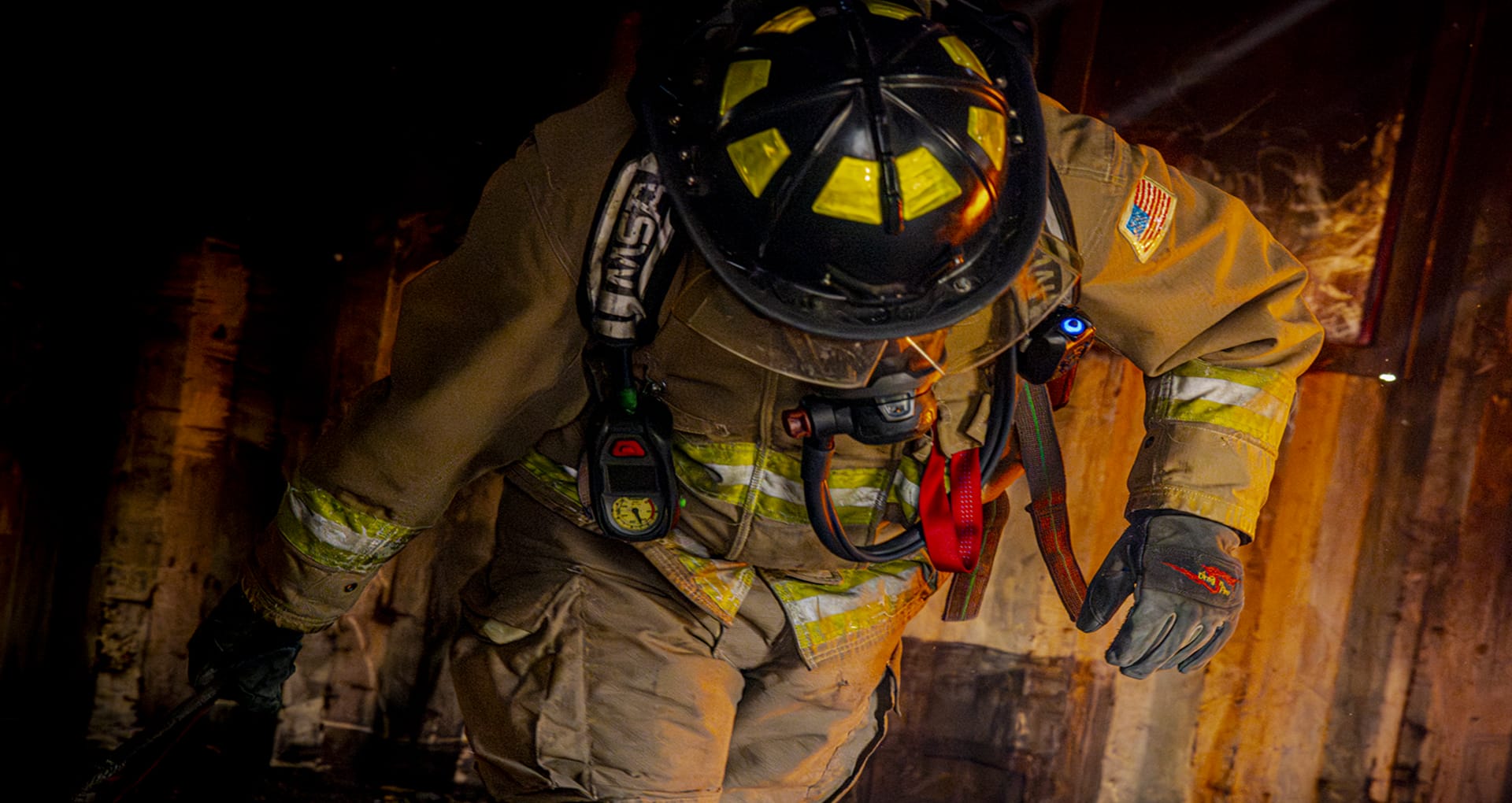 Firefighter in full gear walking towards the camera, bent over as if pulling a weight. The glow of a fire and flames are visible behind him