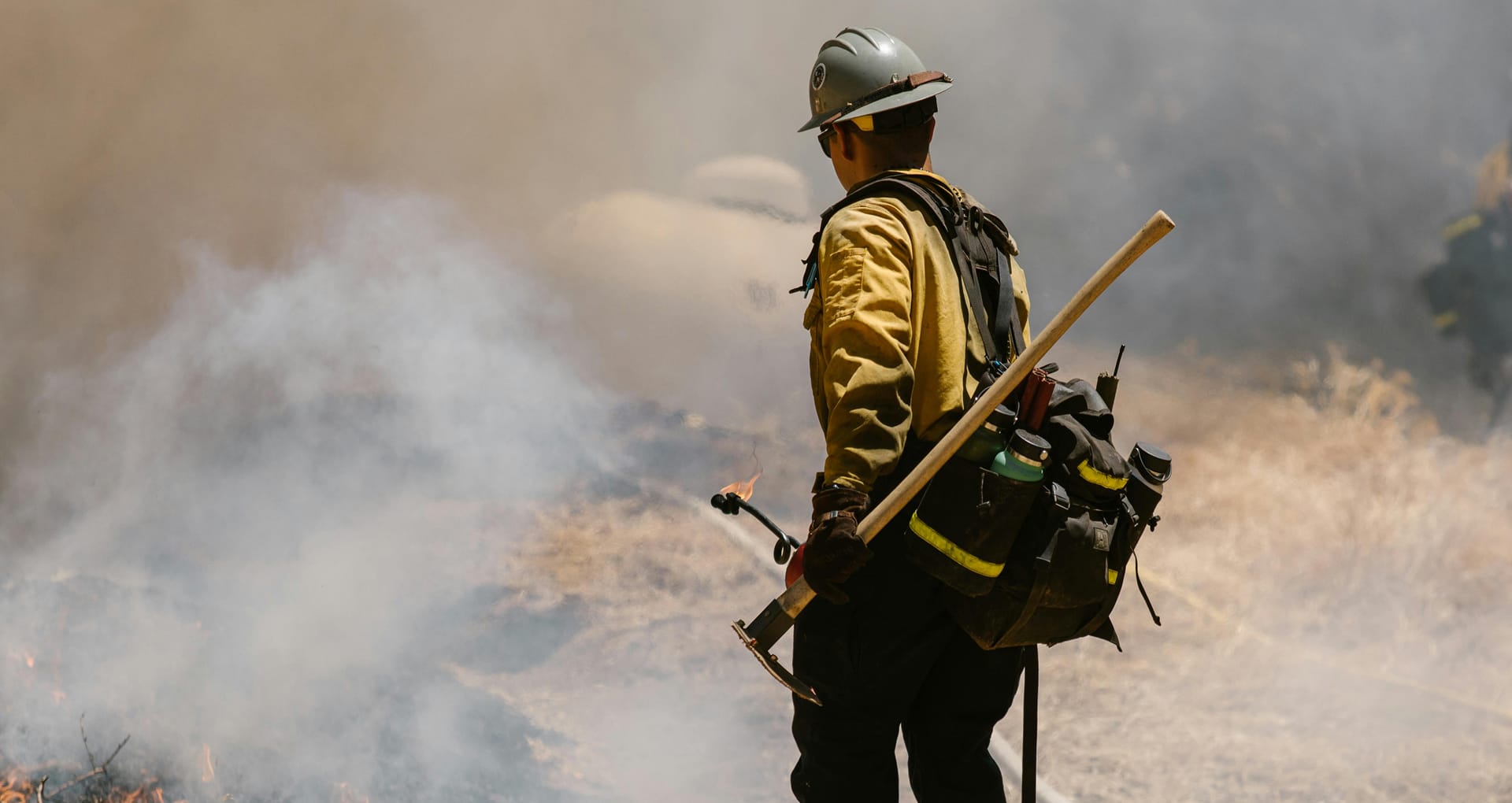 A firefighter outside, wearing a helmet and holding a fighter mans hoe and drip torch, watching a small brushfire in front of him
