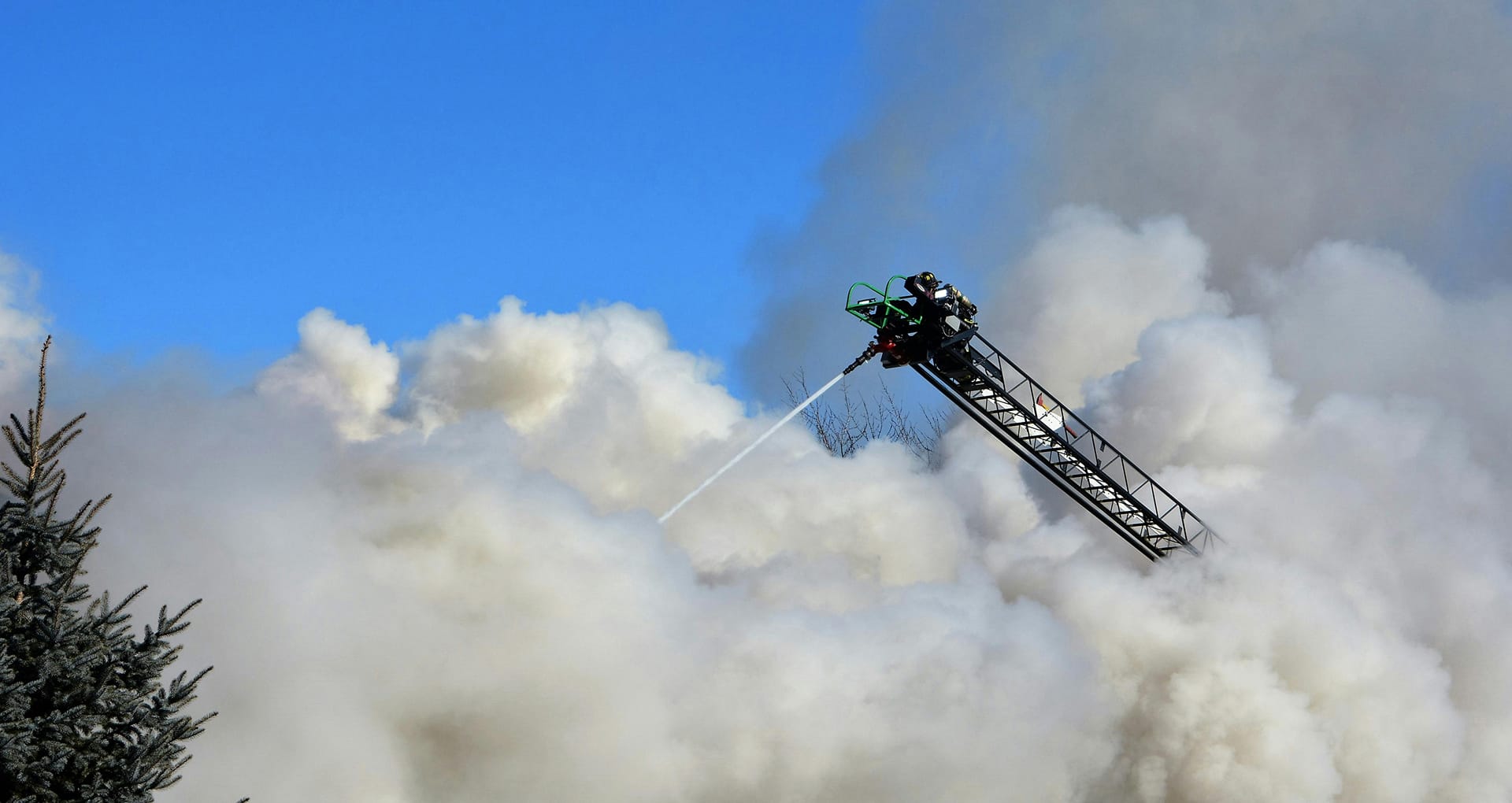 Fireman operating an aerial ladder, with heavy, low laying smoke obscuring what is on fire. Blue sky visible above the smoke, and the tops of trees visible.
