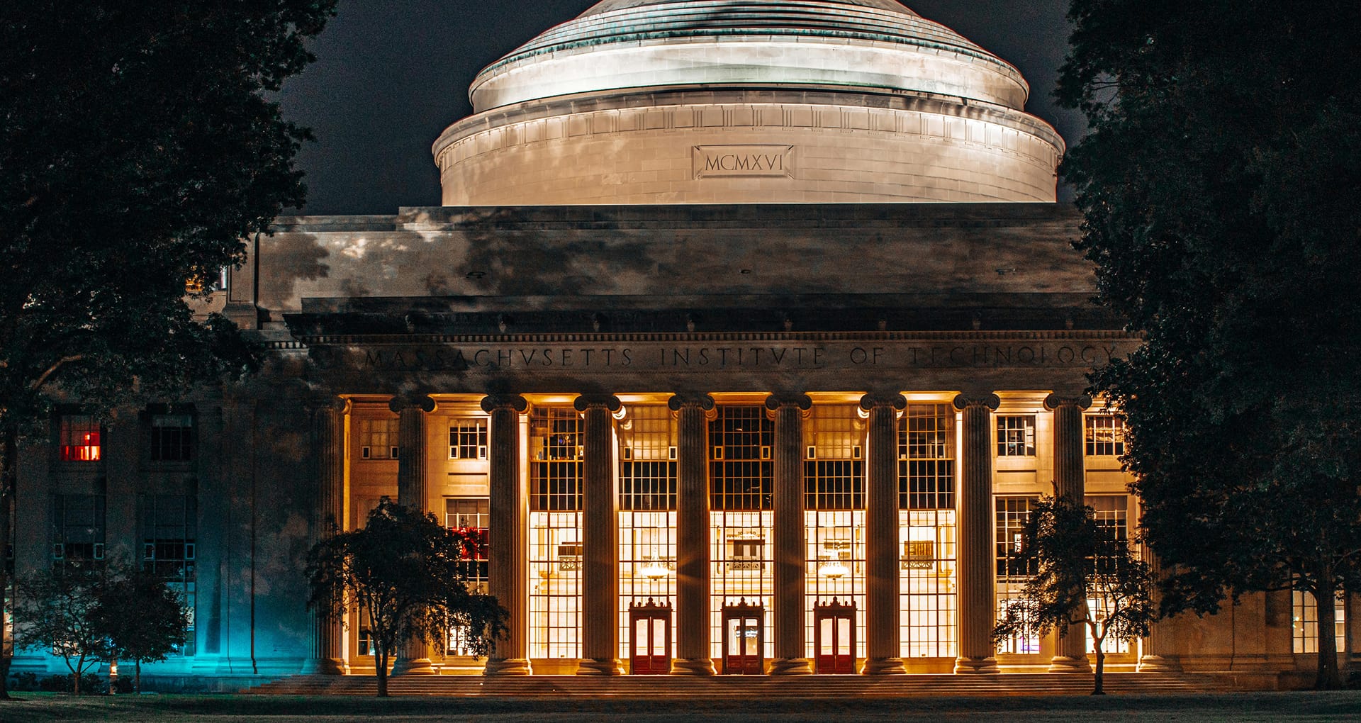 A night time photo of the Massachusetts Institute of Technology, with interior lights illuminating the building, and trees framing the building.