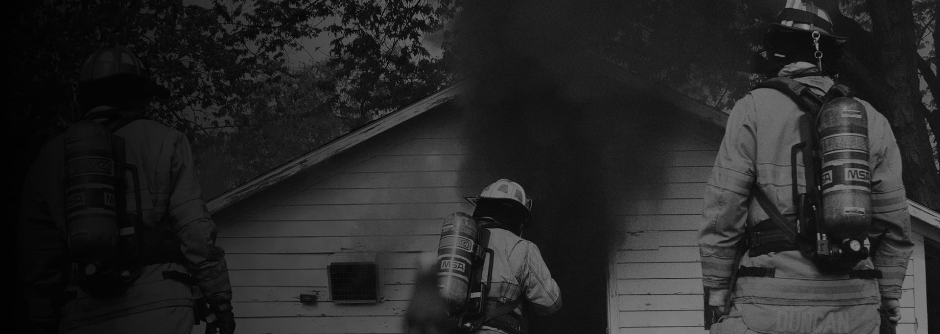 Black-and-white scene of firefighters wearing air tanks approaching a small house, partially obscured by a dark vertical element in the foreground.