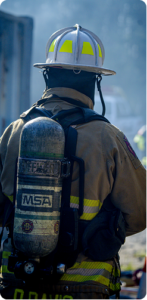 Firefighter seen from behind wearing a helmet and air tank, standing in a smoky scene.