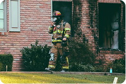 Firefighter in full gear stands outside a brick house with smoke damage around a window, holding a tool on the lawn.