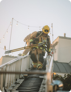 Firefighter climbs an extended ladder toward a roof, carrying gear, with string lights visible overhead.