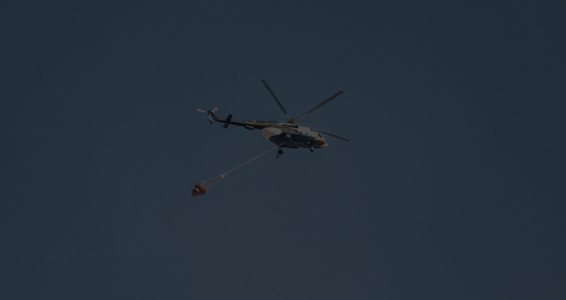 Firefighting helicopter carrying a water bucket in flight against a dark overcast sky