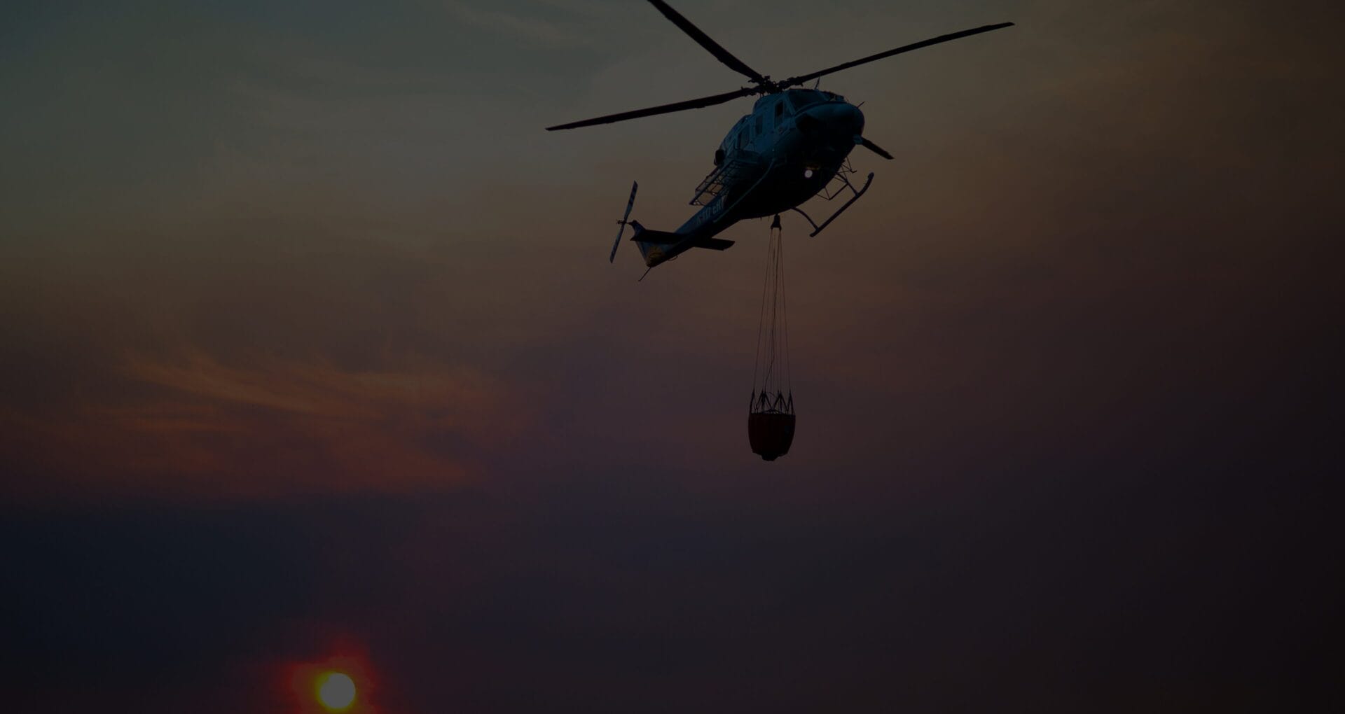 Silhouette of a firefighting helicopter carrying a water bucket against a sunset sky
