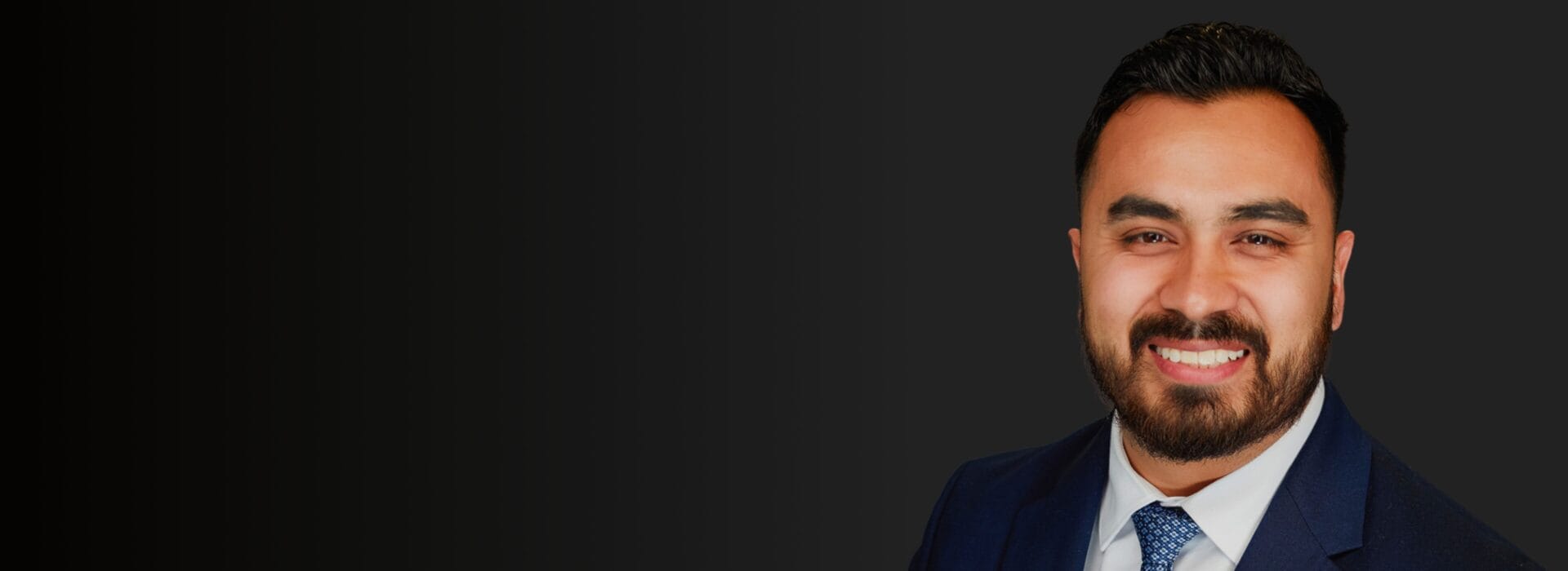 Professional headshot of David Flores in a navy suit and blue tie smiling against a dark background