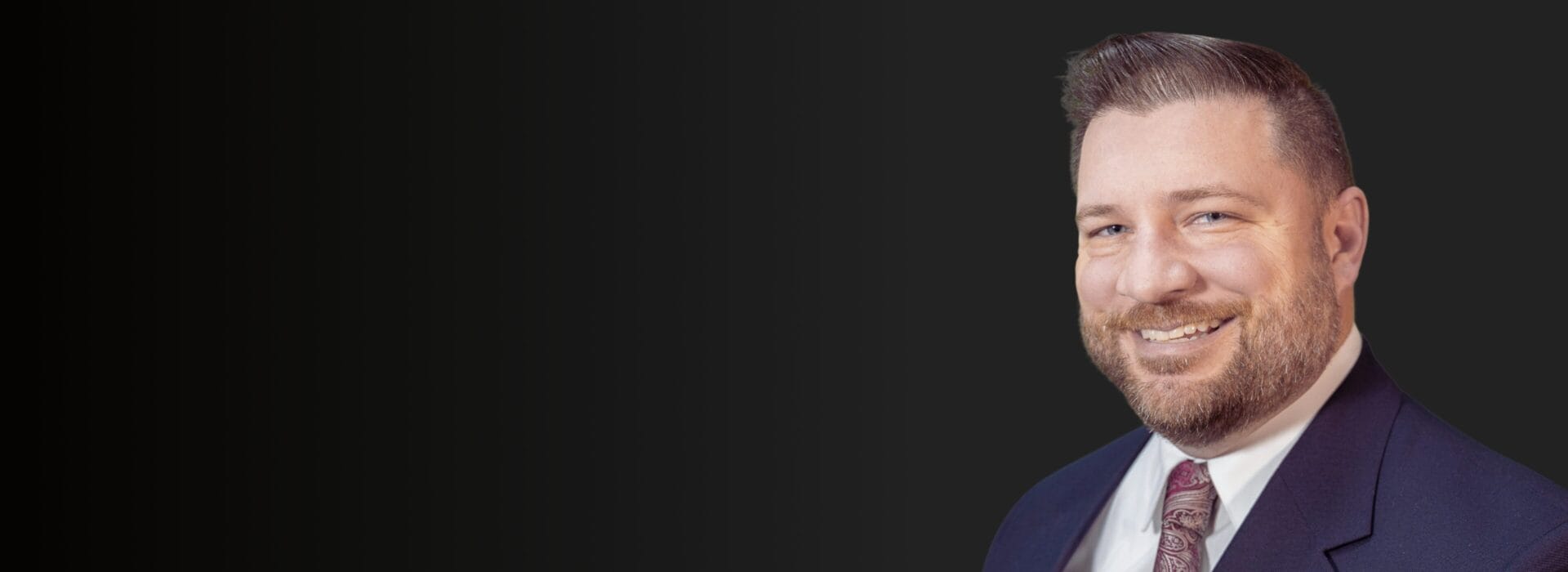 Professional headshot of a Jim Dubina in a navy suit and burgundy paisley tie smiling against a dark background