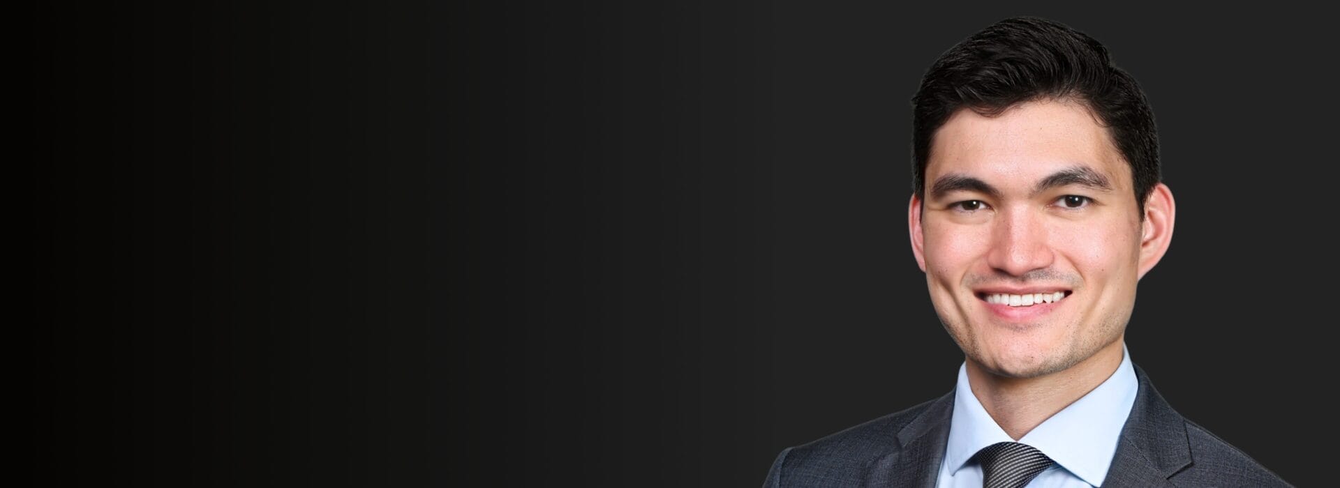 Professional headshot of Roman Moriarty in a charcoal suit and striped tie smiling against a dark background