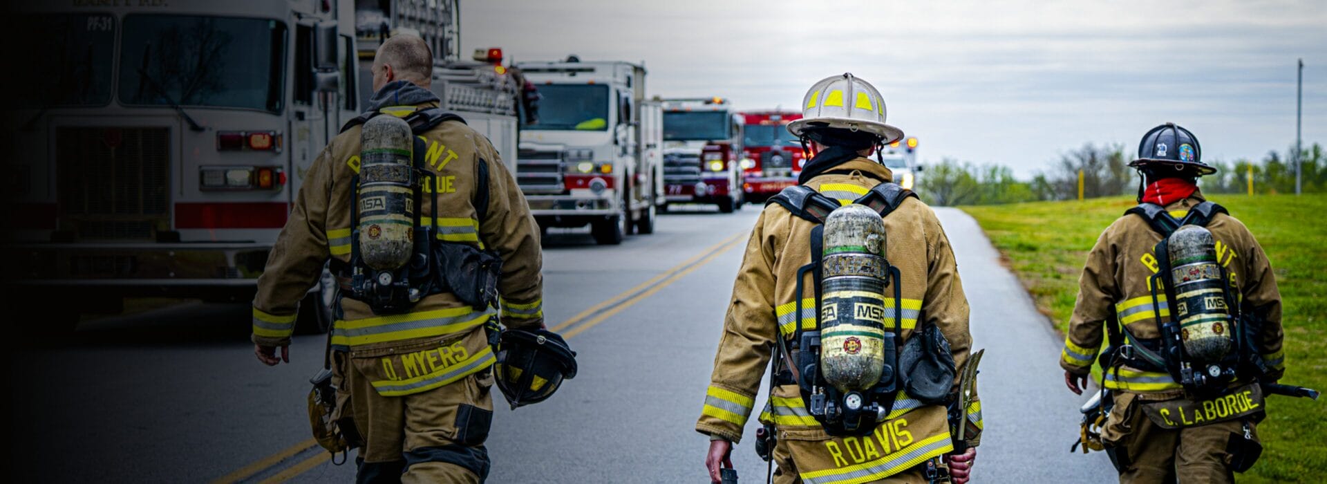 Three firefighters in full gear walking toward a line of fire trucks on a road