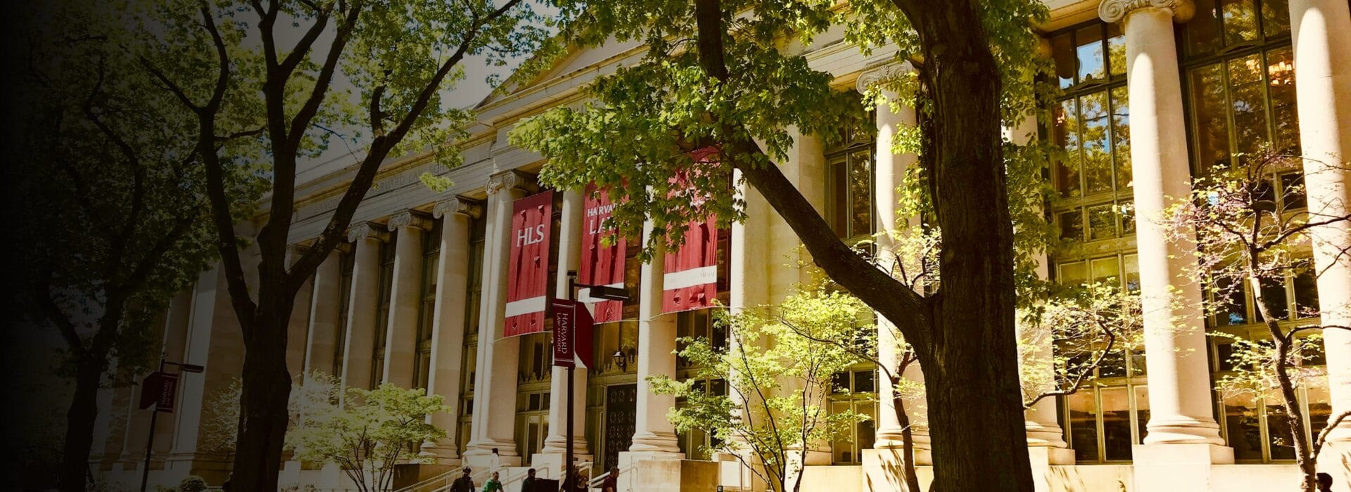 Exterior of Harvard Law School with red HLS banners hanging between white columns, framed by green trees