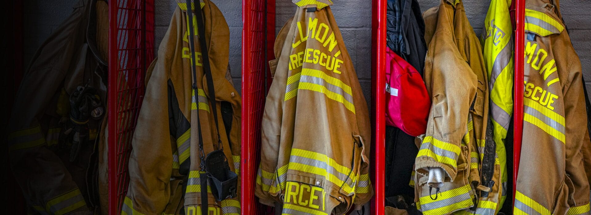 Firefighter turnout gear with yellow reflective strips hanging on red racks in a firehouse