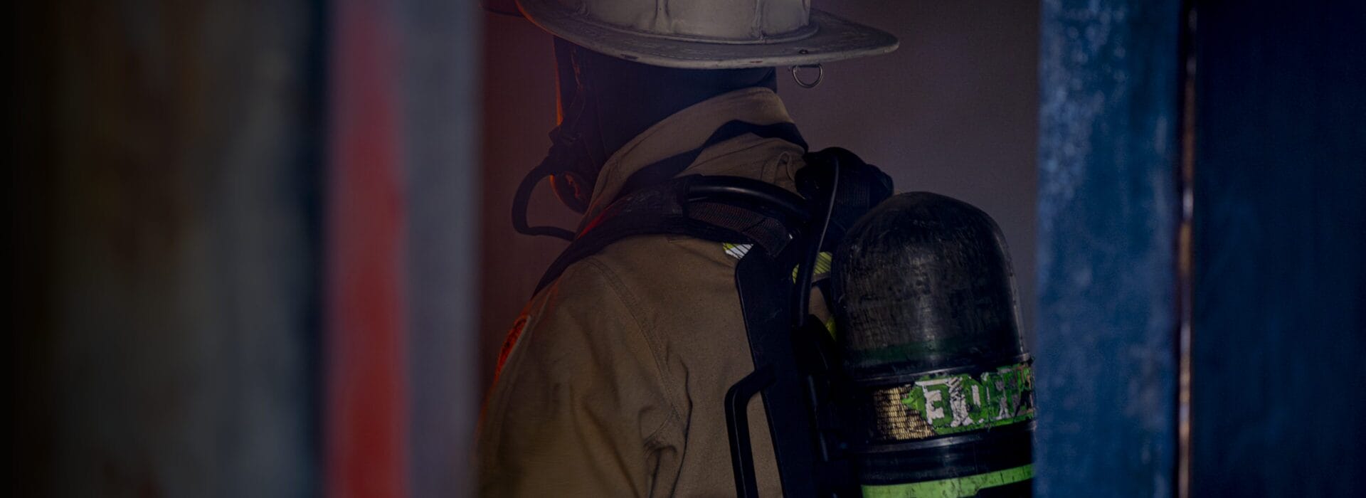 Firefighter in turnout gear and helmet carrying an SCBA tank while approaching a doorway