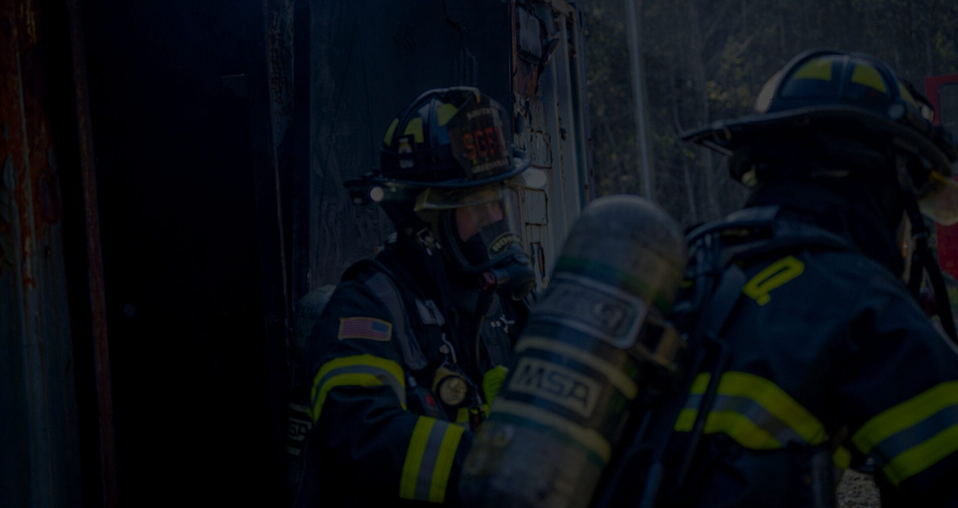 Two firefighters in full gear and face masks working at the entrance of a smoke-filled structure