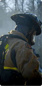 Side profile of a firefighter in turnout gear and helmet standing amid smoke at an outdoor scene
