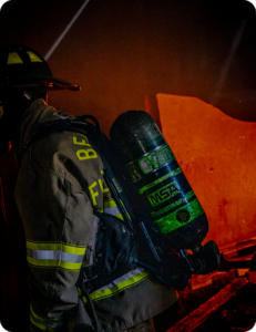 Rear view of a firefighter carrying an SCBA tank while working inside a structure lit by orange firelight