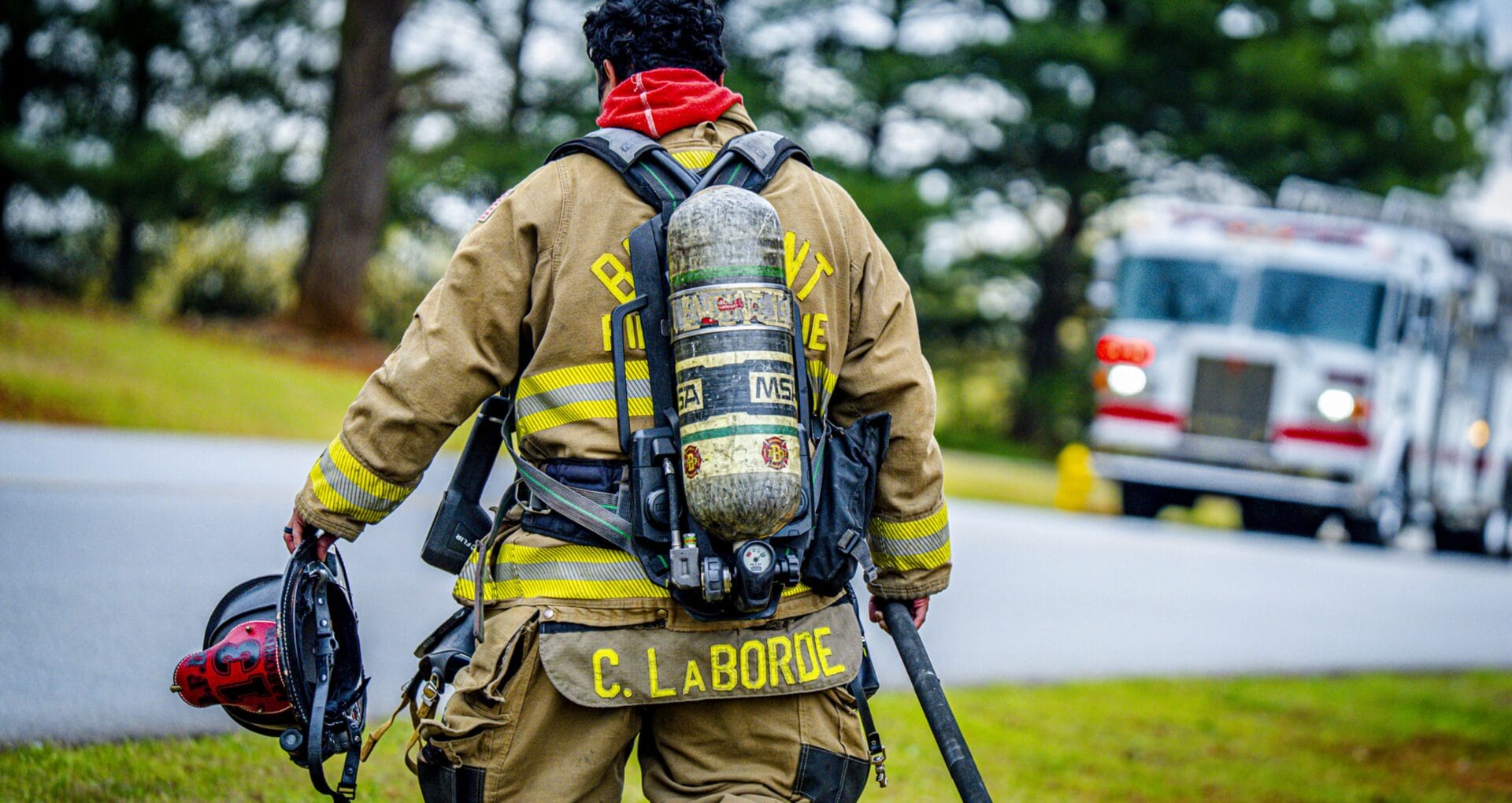 Rear view of a firefighter in full turnout gear carrying a helmet and walking toward a fire truck