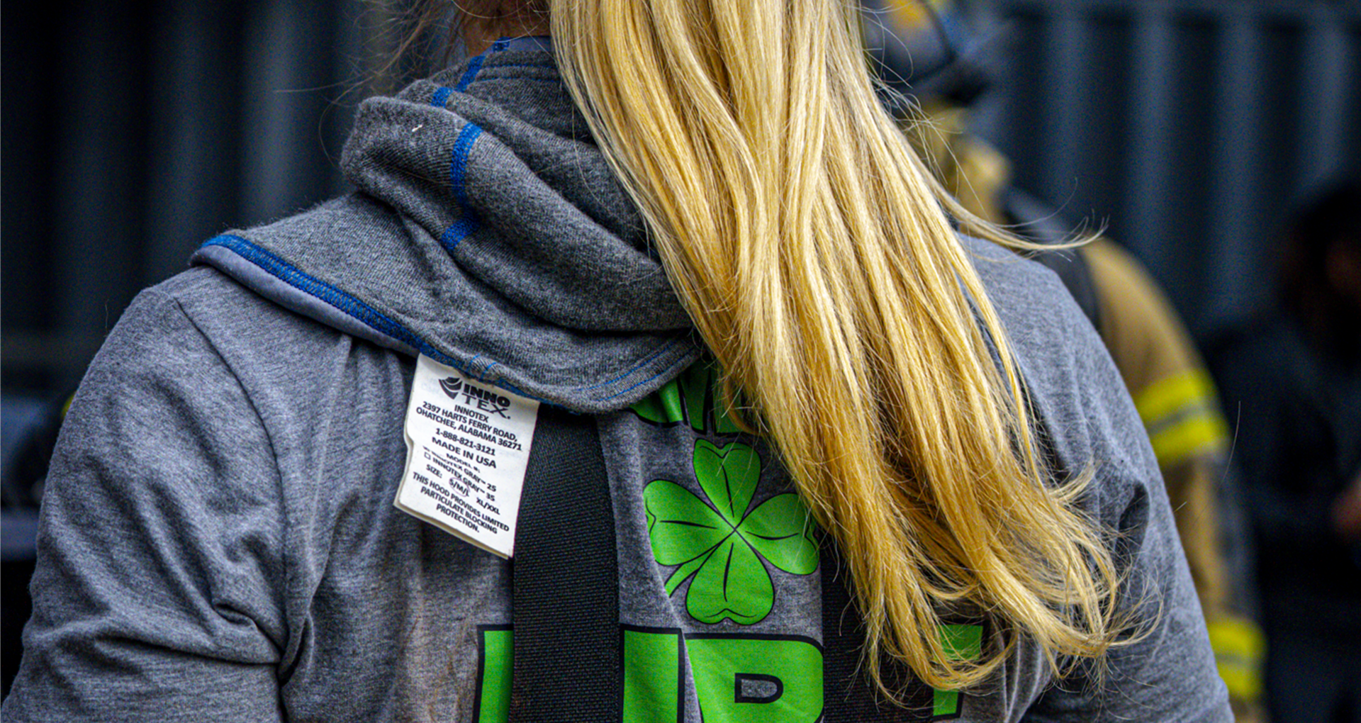 Back view of a firefighter wearing a protective hood and shamrock graphic top at a fire scene