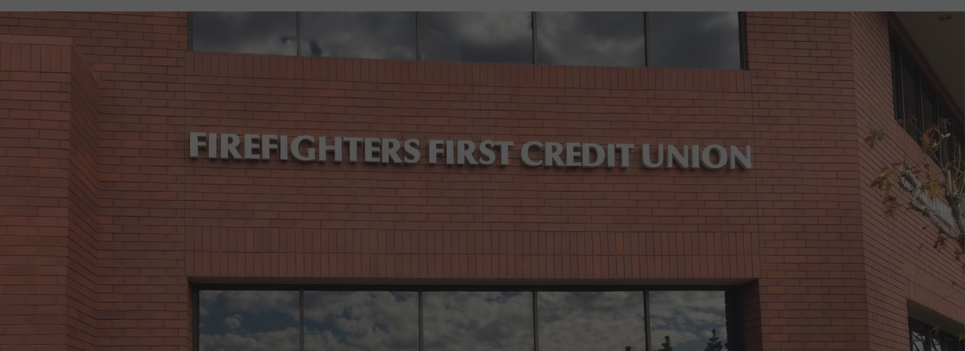 Exterior of a Firefighters First Credit Union branch, featuring a red brick building facade with the credit union name in large letters above a row of reflective windows and a raised brick planter with green shrubs.