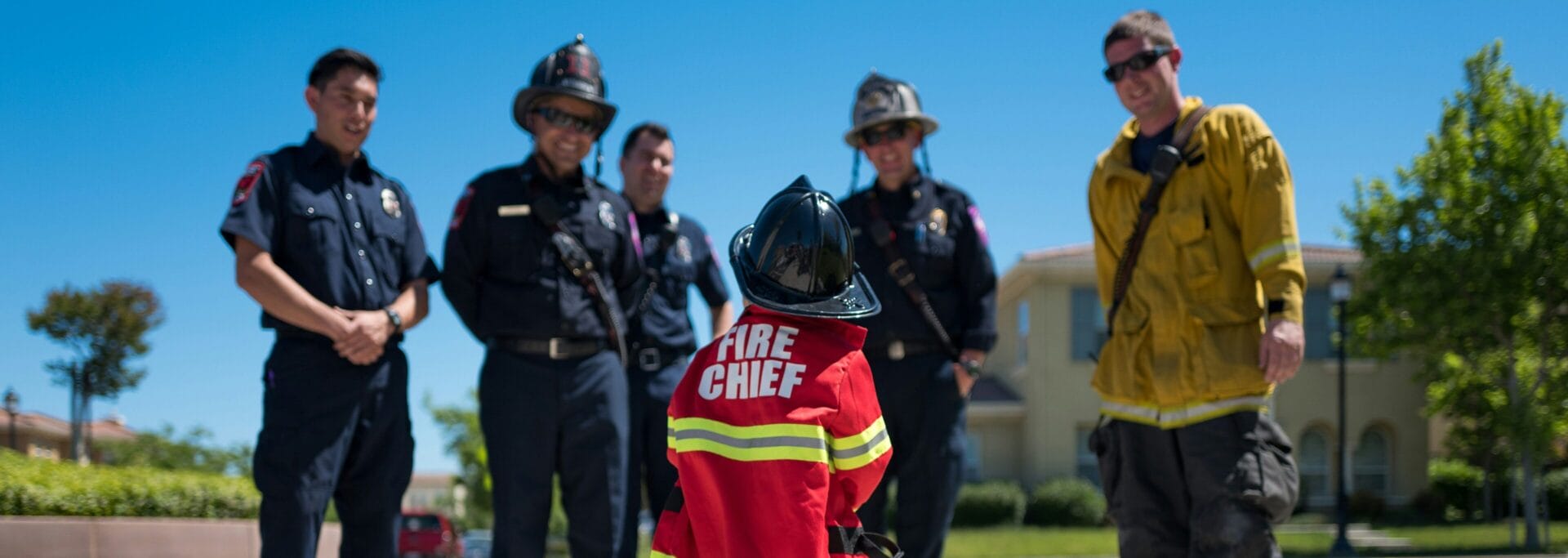 Child in a Fire Chief costume facing a group of first responders and a firefighter outdoors