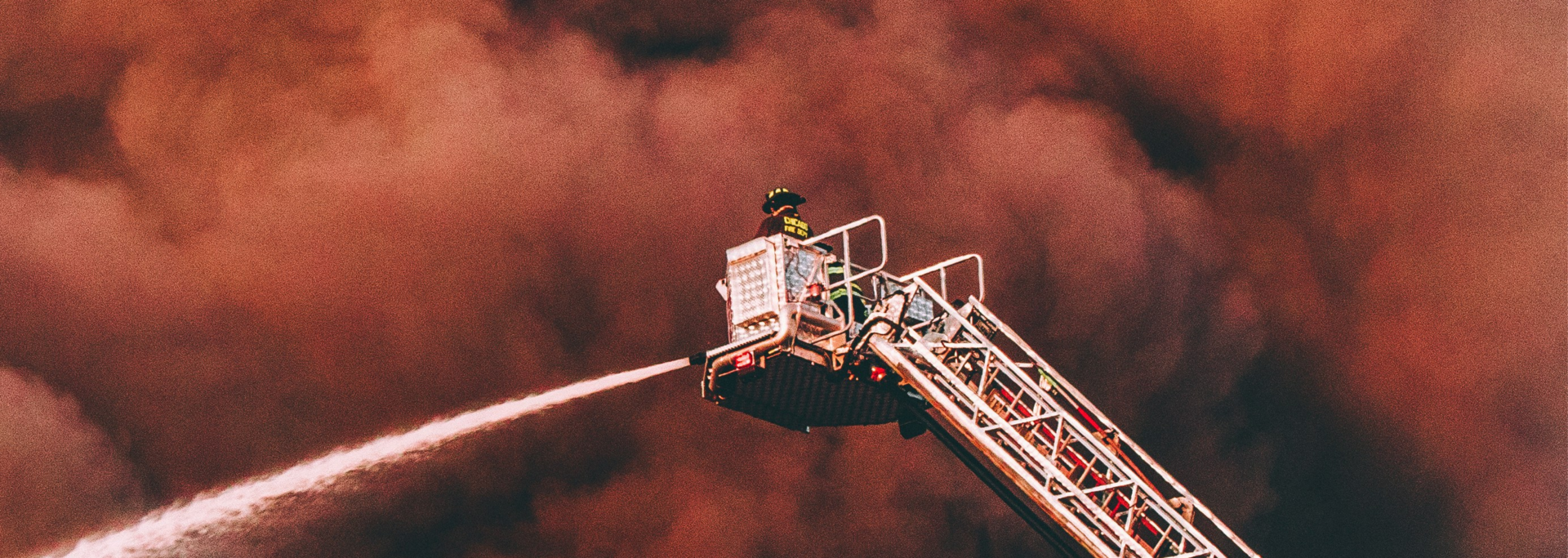 Firefighter in an aerial bucket operating a water hose against a dark, smoke-filled sky